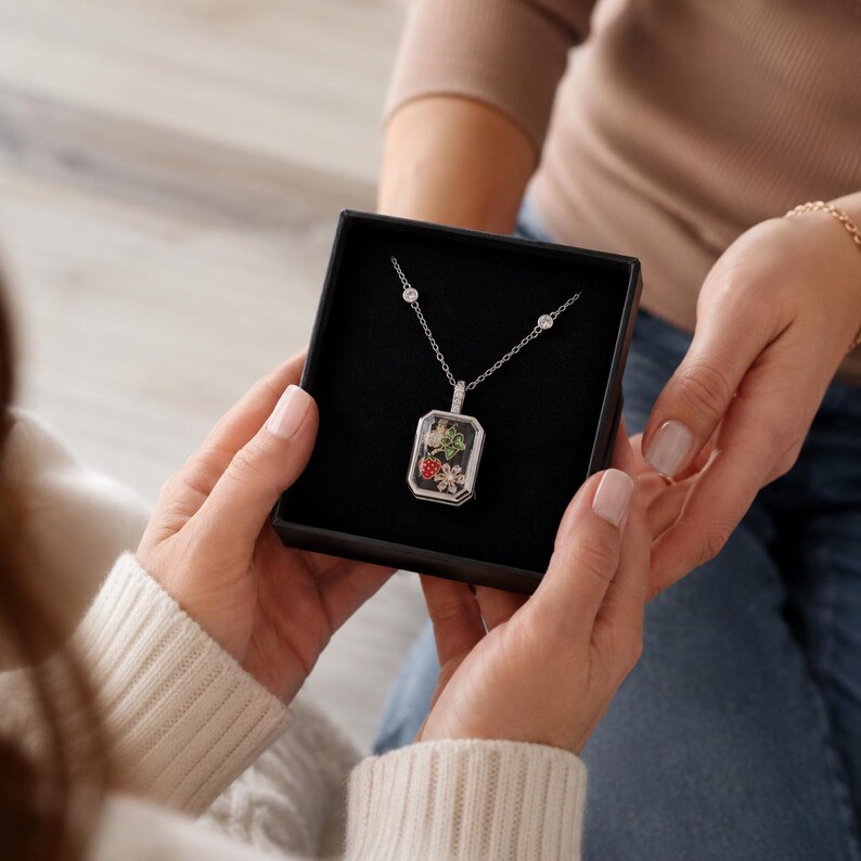 May include: A silver-toned necklace featuring a rectangular pendant with a clear front, containing small colourful charms of a strawberry, a green leaf, and a white flower. The chain includes two small round clear stones set along the silver links.