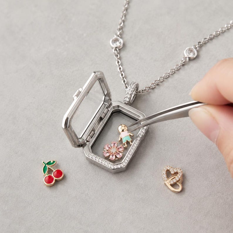 May include: A silver-toned locket necklace with a hinged glass frame containing a small gold-toned figure with a light blue shirt and a pink flower charm. A pair of tweezers holds the figure. Nearby are a red cherry charm and a gold heart charm.