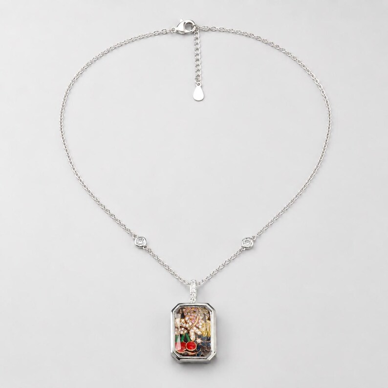 May include: A silver-toned necklace featuring a rectangular pendant filled with small, colourful charms including red cherries, gold-toned shapes, and clear crystals. The chain includes two small circular accents and a lobster clasp closure.