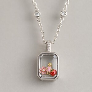 May include: A silver-toned necklace featuring a rectangular pendant with a clear glass front. Inside the pendant are three small charms: a pink flower, a gold-toned figure in a pink dress, and a red strawberry. The chain has two small clear stones.