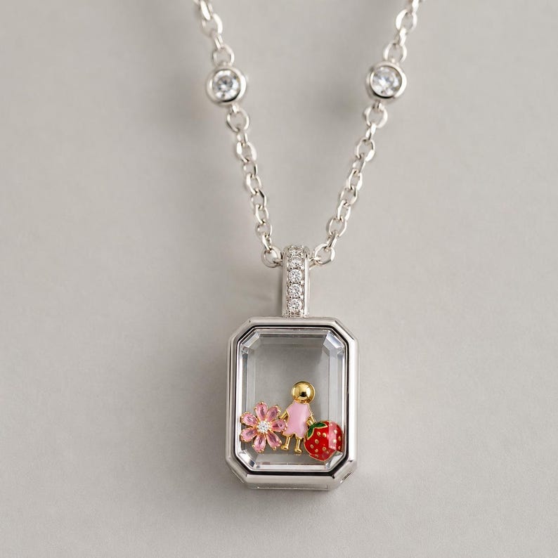 May include: A silver-toned necklace featuring a rectangular pendant with a clear glass front. Inside the pendant are three small charms: a pink flower, a gold-toned figure in a pink dress, and a red strawberry. The chain has two small clear stones.