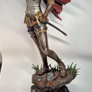 May include: A detailed figurine of a female knight, clad in silver armor and a red cape, holding a sword. The figure stands on a rocky base with green accents, showcasing intricate details and craftsmanship.