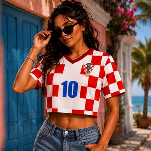 May include: A short-sleeved V-neck crop top featuring a red and white checkered pattern. The front displays the number 10 in blue and a small crest with the letters HNS above a soccer ball graphic. The garment is paired with blue denim jeans.