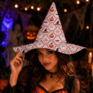 May include: A pointed witch hat featuring a light pink background with a repeating diamond pattern. The print includes small illustrations of orange jack-o'-lanterns, white ghosts, black bats, spiderwebs, and pink bows. The brim has a black underside.