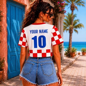 May include: A white cropped t-shirt featuring a red and white checkered pattern on the sleeves and bottom hem. The back of the shirt displays the blue text "YOUR NAME" above the large blue number "10". The shirt is paired with blue denim shorts.