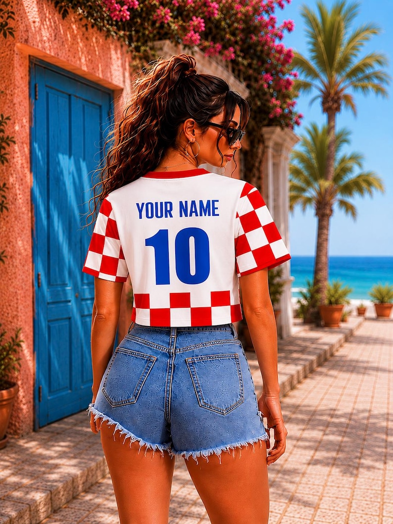 May include: A white cropped t-shirt featuring a red and white checkered pattern on the sleeves and bottom hem. The back of the shirt displays the blue text "YOUR NAME" above the large blue number "10". The shirt is paired with blue denim shorts.