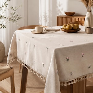 May include: A beige rectangular tablecloth featuring a delicate embroidered pattern of small pink and green floral sprigs. The edges of the fabric are finished with a decorative fringe trim, creating a textured border around the entire perimeter.