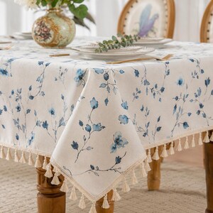 Op de afbeelding: Een rechthoekig tafelkleed van crèmekleurige stof met een delicaat blauw bloemenpatroon van stengels en blaadjes. De randen van het tafelkleed zijn afgewerkt met een decoratieve franje van kleine, lichtbeige kwastjes die naar beneden hangen.