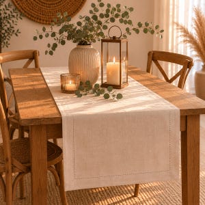 Peut inclure: Un chemin de table en lin beige avec une bordure cousue, posé sur une table en bois. La table est ornée d'un vase de verdure, d'une bougie dans un photophore et d'une lanterne avec une bougie allumée. L'ambiance est chaleureuse et rustique.