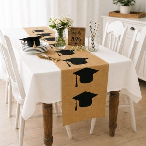 Op de afbeelding: Een tafelsetting met een graduatiethema, met een wit tafelkleed en een jute loper bedrukt met zwarte afstudeerpetten. Een kleine afstudeerpet staat op een stapel witte borden. Een houten bordje leest "Congrats 2026".