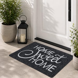 以下が含まれることがあります： 白い筆記体で「HOME Sweet HOME」と書かれた濃い灰色のドアマット。 マットは長方形で、白いドアの前に置かれています。 ろうそくの入った黒いランタンと鉢植えの植物が近くにあります。