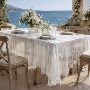 May include: A rectangular dining table covered with a white lace tablecloth featuring intricate floral patterns and scalloped edges. The table is set with white plates, clear glassware, and a large centerpiece of white roses in a glass vase.