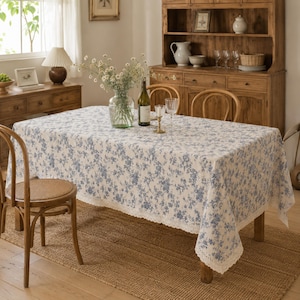 Puede incluir: Un mantel rectangular con un estampado floral en azul y blanco que presenta un delicado borde de encaje de ganchillo. La tela cubre una mesa de comedor de madera, creando una estética suave con su intrincado diseño de estampado botánico.