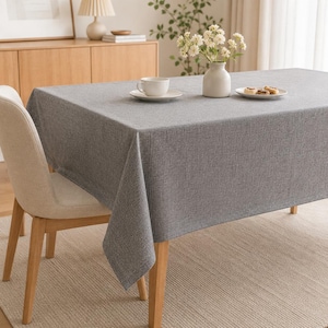 Op de afbeelding: Een rechthoekige eettafel bedekt met een grijs tafelkleed van gestructureerde stof. Op de tafel staat een witte keramische kop op een schoteltje, een witte keramische vaas met witte bloemen en een klein wit bordje met twee gebakjes.