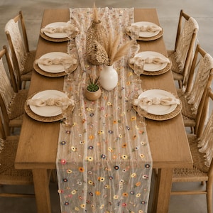 Könnte beinhalten: Ein Holztisch, gedeckt für ein Essen. Der Tisch ist mit einem floral bestickten Tischläufer, gewebten Platzsets, weißen Tellern und beigen Servietten geschmückt. Gewebte Stühle umgeben den Tisch, und eine Vase mit Trockenblumen und einer Sukkulente ergänzen die Dekoration.