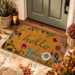 Könnte beinhalten: Eine rechteckige Fußmatte mit beigem Hintergrund und floralem Rand. Das Wort "hello" ist in grüner Schrift in der Mitte geschrieben. Der florale Rand umfasst rote Mohnblumen, blaue Vergissmeinnicht und andere bunte Blumen.