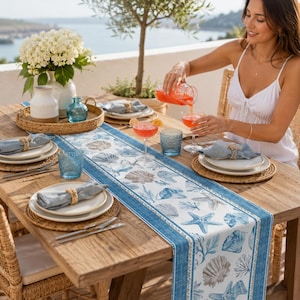 Puede incluir: Un camino de mesa rectangular con un estampado costero en azul y blanco de conchas, estrellas de mar y coral. El camino está colocado sobre una mesa de madera con manteles individuales trenzados, platos blancos y cristalería azul.