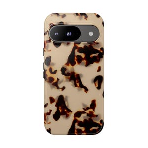 May include: A phone case with a tortoiseshell pattern. The case has a tan background with dark brown irregular shapes. The camera lens is visible at the top, and the case has a rounded rectangular shape.