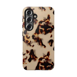 May include: A phone case with a tortoiseshell pattern. The case has a light beige background with irregular dark brown shapes. The phone case has a camera module with three lenses.