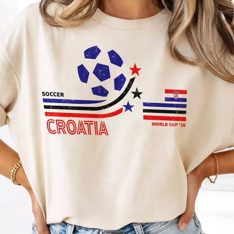 Peut inclure: Un t-shirt couleur cr&egrave;me arborant un motif compos&eacute; d'un ballon de football bleu, de trois &eacute;toiles rouge, noire et bleue, et de rayures horizontales. Le texte indique SOCCER CROATIA et WORLD CUP '26, avec un petit drapeau croate.
