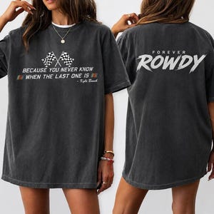 Op de afbeelding: Donkergrijs t-shirt met de tekst "BECAUSE YOU NEVER KNOW WHEN THE LAST ONE IS" en twee geblokte vlaggen. De achterkant van het shirt zegt "FOREVER ROWDY" in het wit. Een casual unisex t-shirt.