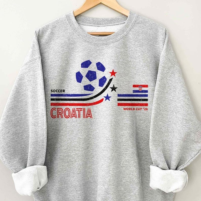Peut inclure: Un sweat-shirt gris clair &agrave; col rond avec un imprim&eacute; graphique repr&eacute;sentant un ballon de football bleu, trois &eacute;toiles et des rayures horizontales bleues, noires et rouges. Le texte indique SOCCER, CROATIA et WORLD CUP '26 avec un drapeau croate.