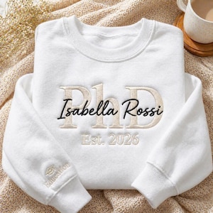 May include: A white sweatshirt with the name "Isabella Rossi" embroidered in black script below the embroidered letters "PhD" in a light tan color. The year "Est. 2026" is embroidered below the name. The left sleeve has a small embroidered graduation cap and the word "Education".