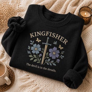 Può includere: Felpa nera con la scritta "KINGFISHER" ricamata in crema, insieme a una spada, fiori e farfalle. Anche la frase "The devil is in the details" è ricamata sulla felpa.