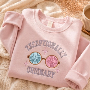 May include: A light pink sweatshirt with the words "EXCEPTIONALLY ORDINARY" embroidered in purple. The design includes glasses with blue and pink spiral lenses. The sweatshirt is folded on a textured surface.