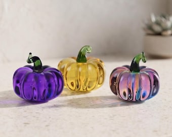May include: Three small glass pumpkins with ribbed surfaces and dark green stems. The left pumpkin is deep purple, the center pumpkin is translucent yellow, and the right pumpkin features an iridescent finish with pink, blue, and purple tones.