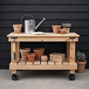 Puede incluir: Un banco de trabajo de jardinería de madera con un estante inferior y una superficie superior. Cuenta con una regadera de metal, macetas de terracota de varios tamaños y una pequeña paleta. El banco está sobre ruedas y sobre un fondo oscuro.
