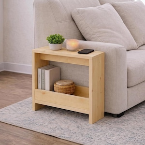Puede incluir: Una mesa auxiliar de madera clara con un estante, que contiene libros y una cesta tejida. Una pequeña planta en maceta, una vela encendida en un vaso y un teléfono inteligente están encima. La mesa está al lado de un sofá beige.