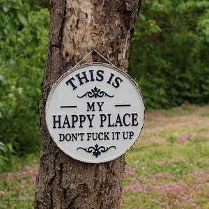 May include: A round, white metal sign with the text "THIS IS MY HAPPY PLACE DON'T FUCK IT UP" in navy blue lettering. The sign is hanging on a tree trunk with a natural background.