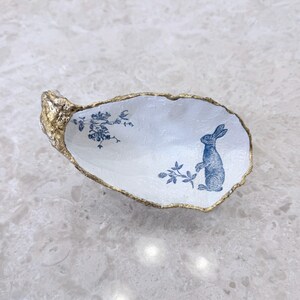 May include: An oval trinket dish, white with a gold-painted rim. The interior has a blue illustration of a rabbit and floral details. The dish is shaped like an oyster shell.