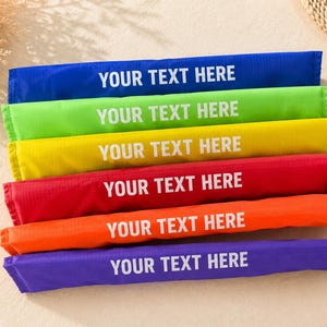 Könnte beinhalten: Sechs farbige, rechteckige Stoffstücke, übereinander gestapelt. Jedes Stück hat eine andere Farbe: blau, grün, gelb, rot, orange und lila. Der Text "YOUR TEXT HERE" ist in Weiß auf jedem Stück gedruckt.