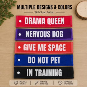 Può includere: Cinque fasce rettangolari in tessuto per guinzagli con bottoni a pressione bianchi. I colori sono rosso, viola, rosso, blu e nero. Ognuna presenta testo bianco: DRAMA QUEEN, NERVOUS DOG, GIVE ME SPACE, DO NOT PET e IN TRAINING. Testo in alto: MULTIPLE DESIGNS & COLORS.