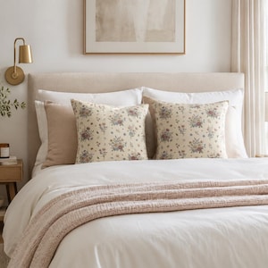 Puede incluir: Dos cojines cuadrados con una base de color crema y un patrón floral repetitivo en tonos apagados de rojo, azul y verde. Los cojines están dispuestos sobre una cama con ropa de cama blanca, una manta texturizada de color rosa claro y un cabecero beige.