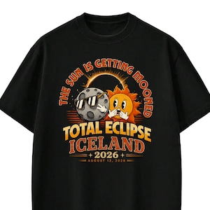 May include: A black t-shirt featuring a graphic of a moon wearing sunglasses and a sun with a surprised expression. The text reads THE SUN IS GETTING MOONED, TOTAL ECLIPSE ICELAND 2026, and AUGUST 12, 2026, with a solar eclipse illustration above.