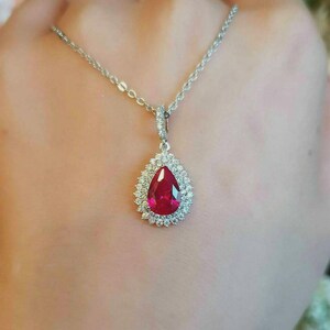 May include: A silver necklace featuring a teardrop-shaped, deep red gemstone pendant. The pendant is surrounded by a halo of small, clear gemstones. The necklace chain is delicate and silver.