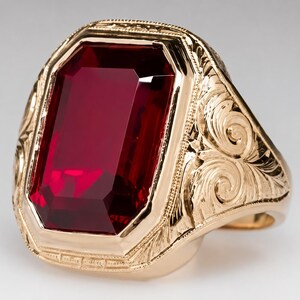 May include: A gold ring with a large, rectangular, faceted, deep red gemstone. The ring has an ornate, engraved design on the band and around the gemstone setting. The ring is set against a neutral background.
