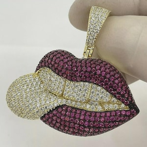 May include: A gold-toned pendant in the shape of lips, encrusted with small, round, ruby-colored and clear gemstones. The pendant has a bail covered in clear gemstones. The lips are in a puckered position.