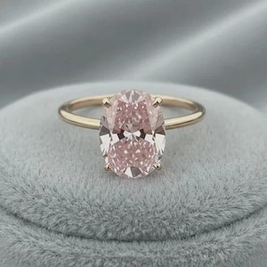 May include: A gold ring featuring an oval-cut, light pink gemstone. The ring has a delicate band and four prongs securing the stone. The gemstone's facets reflect light, creating a sparkling effect. The ring is set against a soft, gray background.