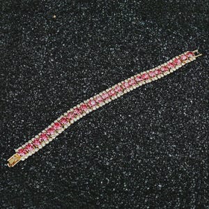 May include: A gold-toned bracelet with a row of pink gemstones and smaller white gemstones. The bracelet is set against a black textured background, showcasing its sparkle and design. The clasp is also gold-toned.