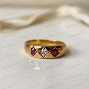 May include: A gold ring featuring a central diamond flanked by two rubies. The ring has a rounded band and the gemstones are set in square settings. The ring is displayed on a white surface.