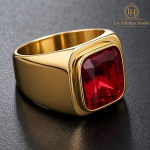 May include: A gold-colored ring with a large, square, red gemstone. The ring has a thick band and a polished finish. The gemstone is set in a square bezel. The ring is displayed on a dark surface. The text "LakshHriday Jewels" is in the upper right corner.