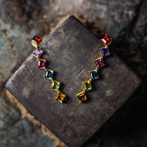 Può includere: Un paio di orecchini dorati con una serie di pietre preziose quadrate e colorate. Le pietre sono disposte a zig-zag, mostrando uno spettro di colori tra cui rosso, viola, rosa, blu, verde e giallo. Gli orecchini sono esposti su una superficie scura e strutturata.