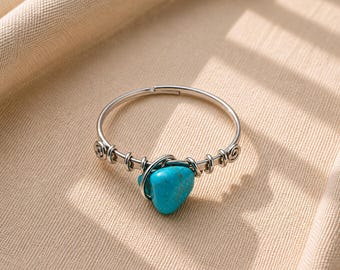Puede incluir: Un anillo de metal plateado con una piedra turquesa brillante de forma irregular, asegurada mediante un alambre enrollado. La banda está hecha de un alambre metálico fino y flexible con detalles en espiral a ambos lados de la piedra.