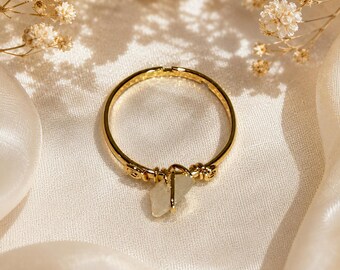 Puede incluir: Un anillo dorado con un pequeño colgante de piedra preciosa de color claro. El anillo se presenta sobre tela blanca, con flores secas en el fondo. El diámetro del anillo es de aproximadamente 2,5 cm.