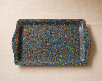 Puede incluir: Una bandeja de servicio rectangular con fondo azul que presenta un patrón denso y repetitivo de pequeñas flores amarillas, flores naranjas y complejos motivos de hojas en rojo y azul oscuro en toda la superficie y bordes.