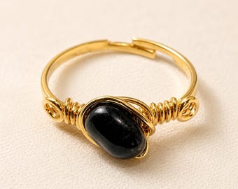 Puede incluir: Un anillo ajustable de tono dorado que presenta una única piedra negra lisa de forma ovalada en el centro. La banda está construida con un hilo fino de color dorado que se enrolla alrededor de la piedra y forma un aro circular abierto.